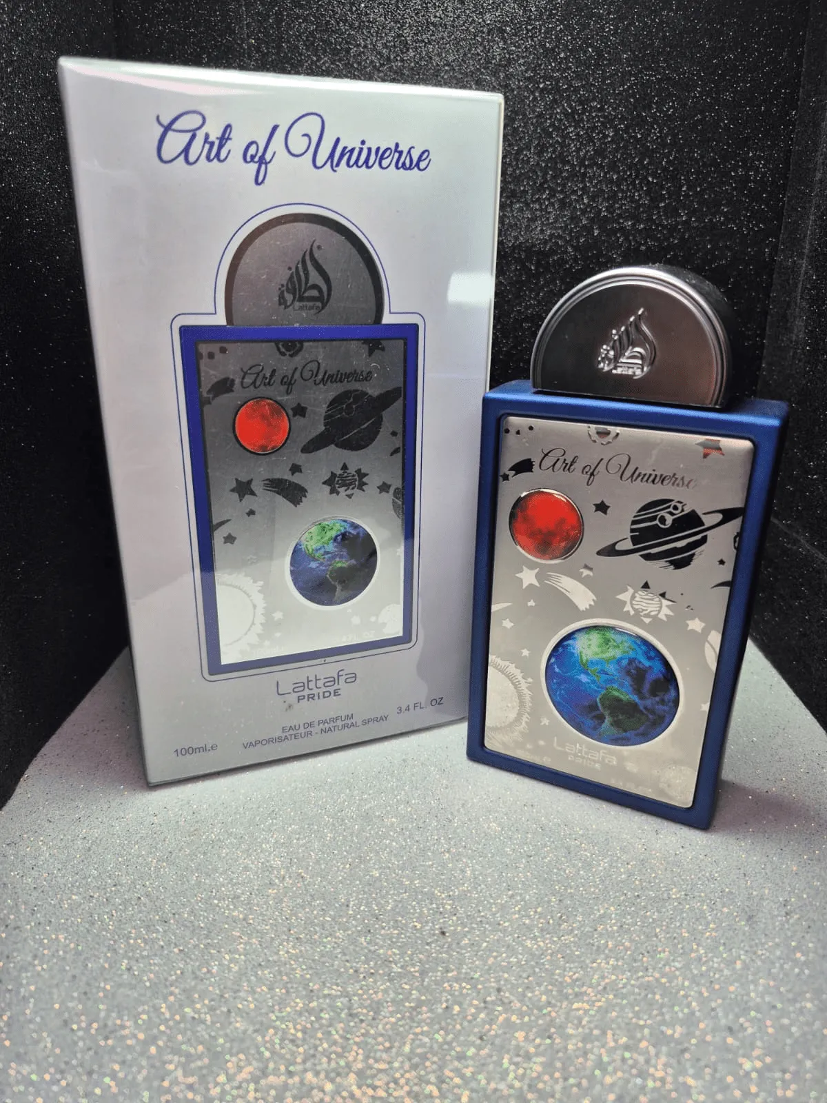 Perfume árabe Out of Universe Lattafa Pride con diseño de galaxia y planetas en el frasco y caja.